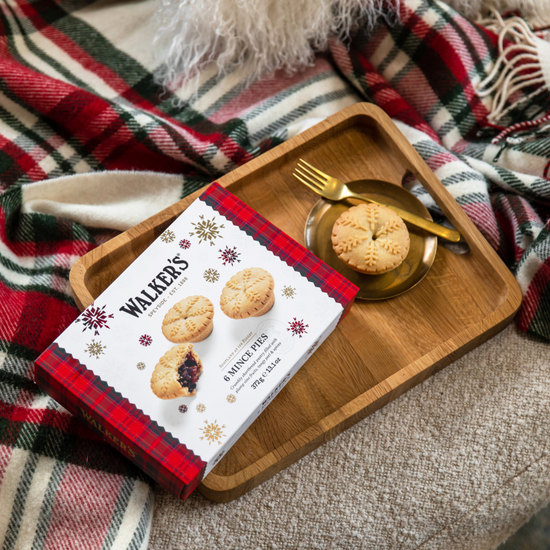 Image of a box of Walker's Mince Pies on a wood tray with a minced pie on a gold plate with fork. Tray is on a couch cushion with plaid blanket.