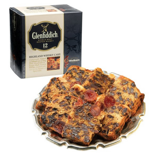 A box of Walker's Glenfiddich Highland Whisky Cake with dark green and cream color packaging in the upper corner with an unboxed Whisky Cake cut in slices on a platter.