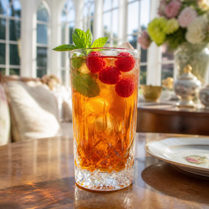 A clear cut-glass tumbler filled with an amber-orange beverage, garnished with fresh mint leaves and three red raspberries, set on a wooden table in a bright, softly blurred indoor setting.