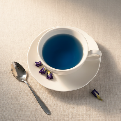 White cup with blue hot tea liquid on a beige surface with a spoon and dried flowers.