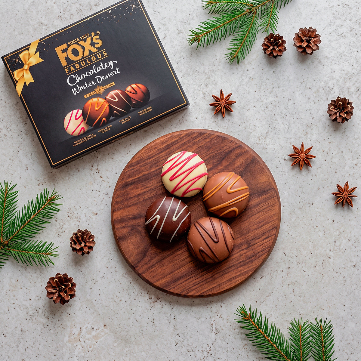Fox’s Chocolatey Centre Cookies box on a white surface with cookies on a wooden board, pinecones, and star anise.