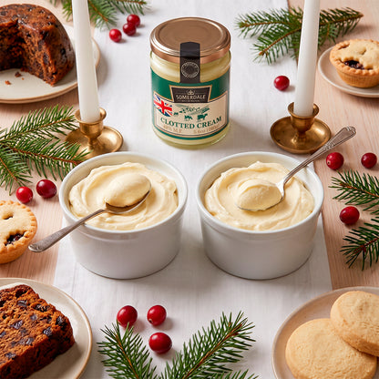 Two bowls of clotted cream on a table with mince pies, cranberries, and pine sprigs