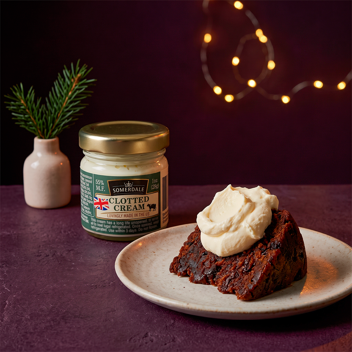 Christmas pudding with clotted cream on a plate next to a jar of cream, festive lights behind