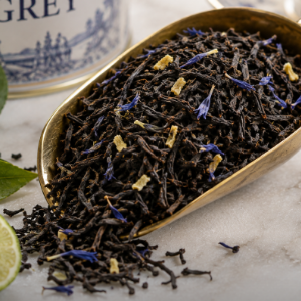 Dark herbal tea loose leaves in a gold scoop, with blue flower petals and small yellow pieces mixed in, set on a light marble surface with a blurred tea canister in the background.