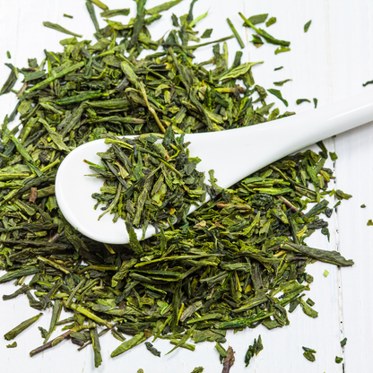 Loose green tea leaves scattered on a white wooden surface with a white ceramic spoon holding some of the leaves.