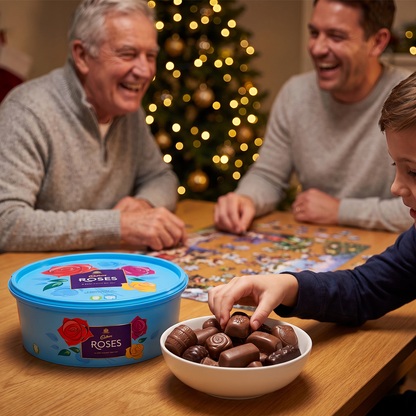 Two men playing a board game with a Roses tub and bowl of chocolates, Christmas tree behind.