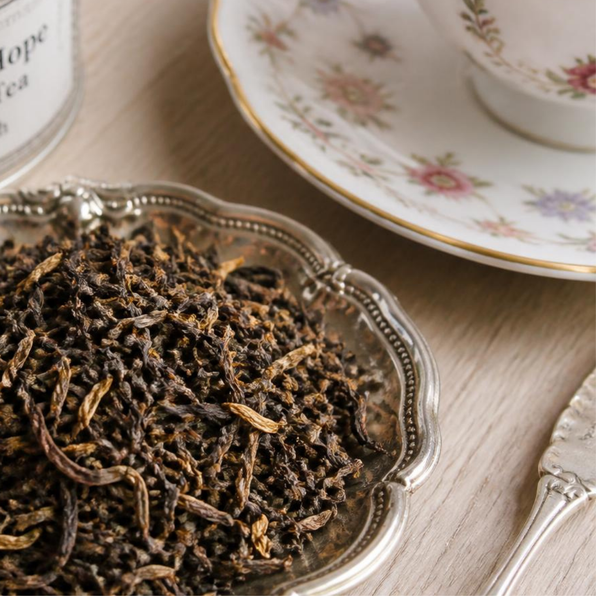 Close-up of loose-leaf Margaret's Hope Darjeeling tea piled in an ornate silver dish on a light wood surface, with a floral teacup and saucer and part of a canister visible in the background.