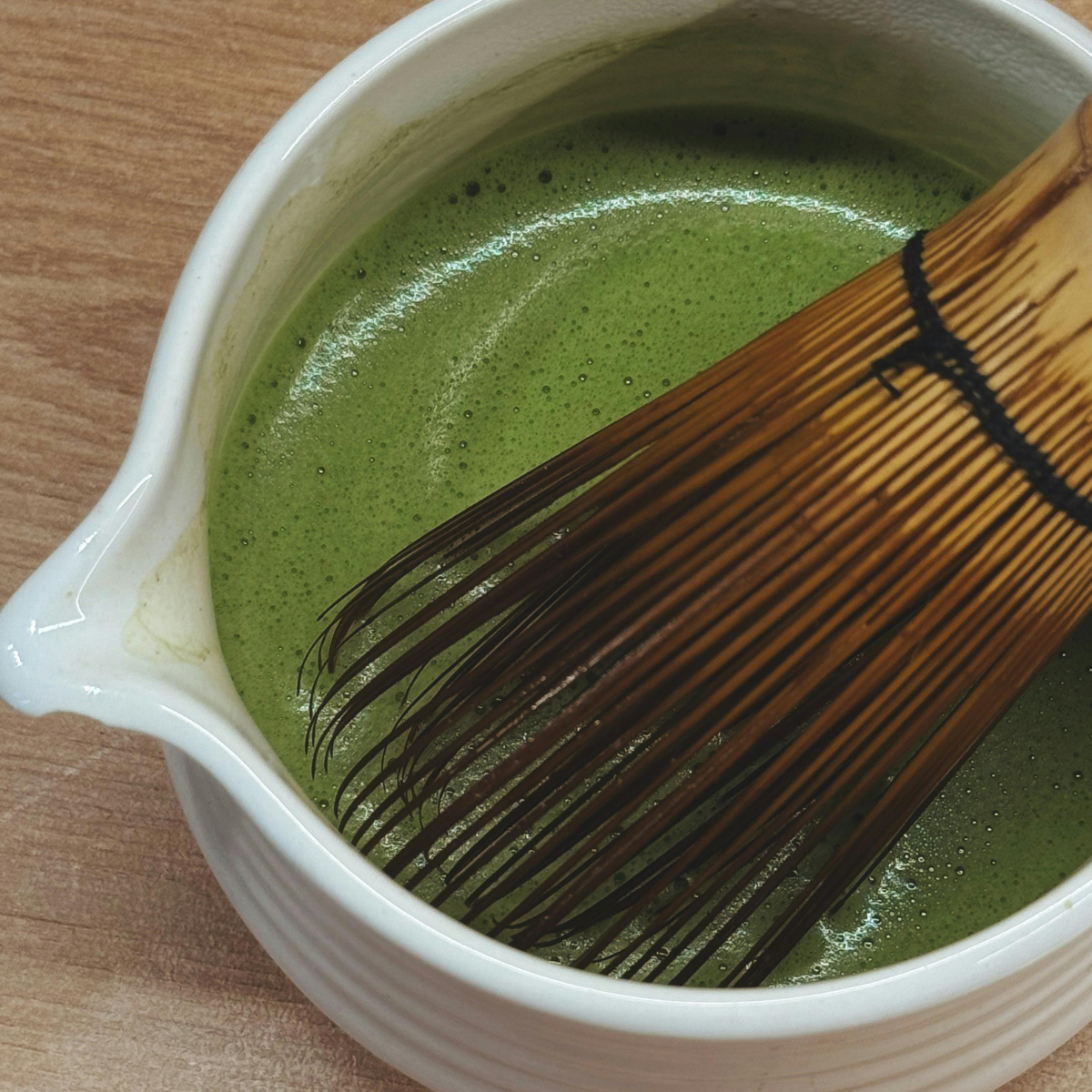 A close-up of green matcha tea being whisked in a white ceramic bowl with a bamboo chashaku whisk resting inside.