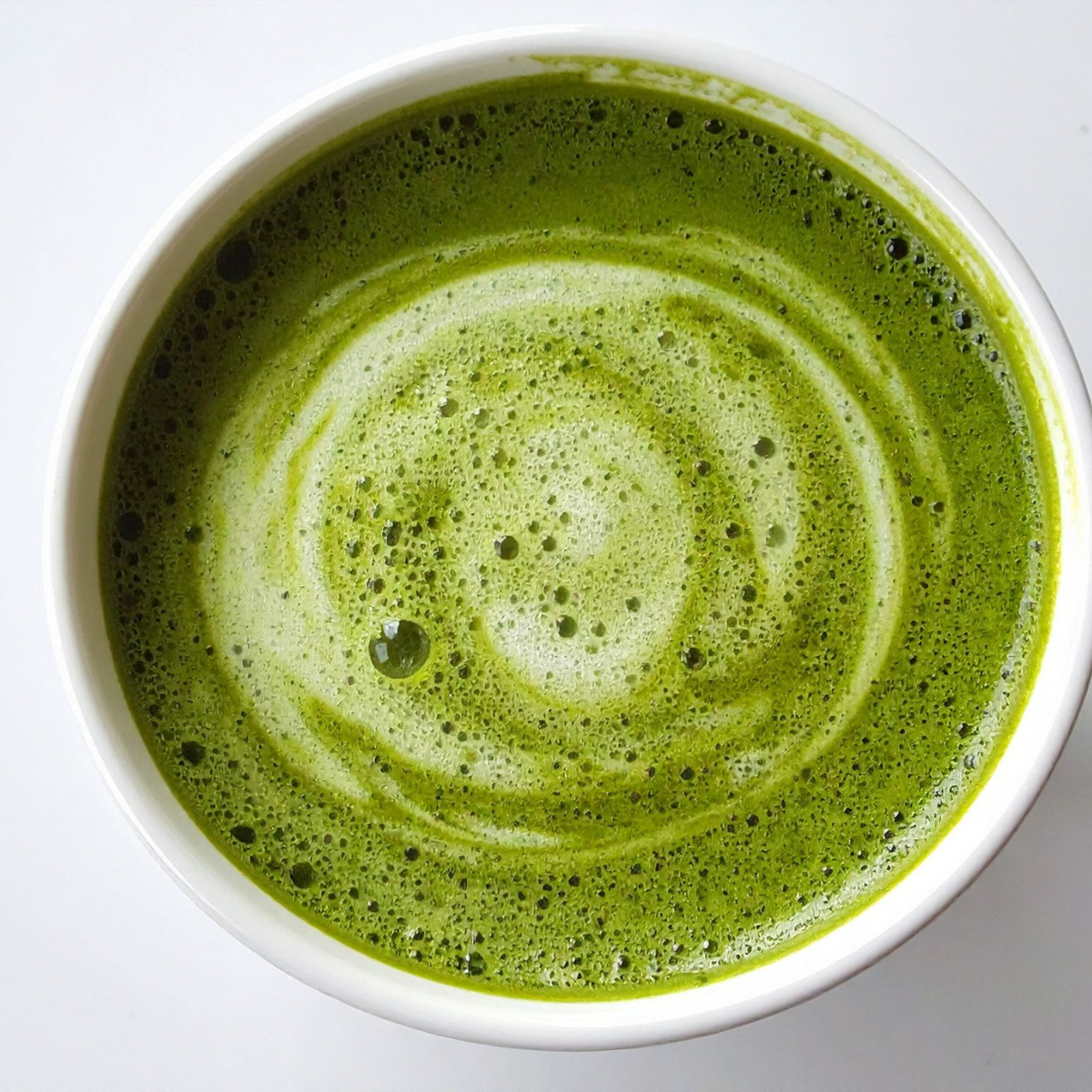 A top-down view of a white ceramic cup filled with a frothy green matcha latte, showing a swirled pattern of foam on the surface.