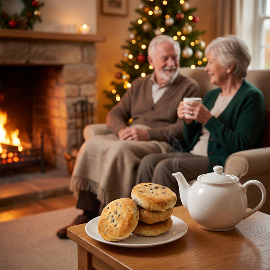 Older couple sitting by a fireplace with a plate of Eccles cakes and a teapot on a table, Christmas tree in the background