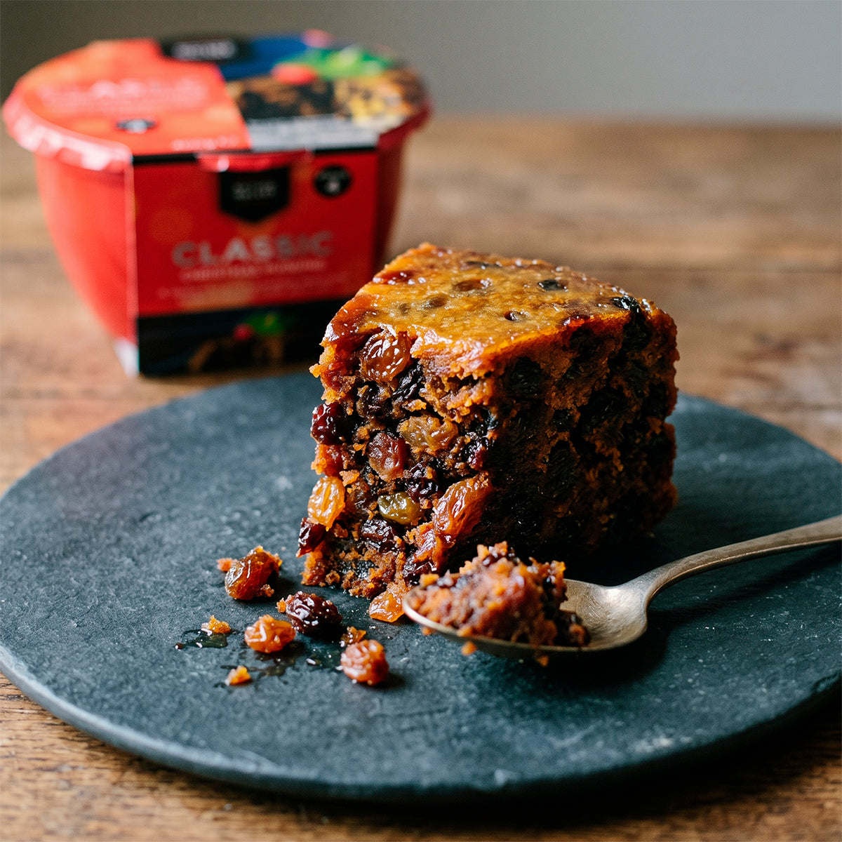 Slice of fruit cake on a slate plate with a spoon, red Clarke’s tub in the background.