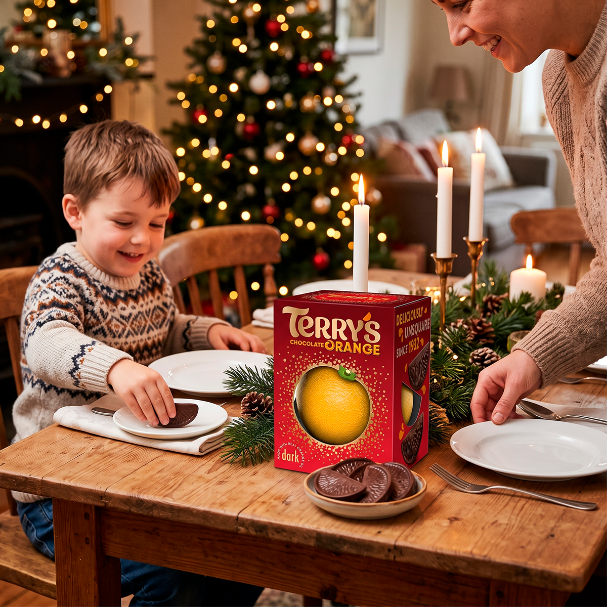 A boy at a table with a Terry’s Chocolate Orange box, a plate of chocolate slices, and lit candles, with a Christmas tree behind him.