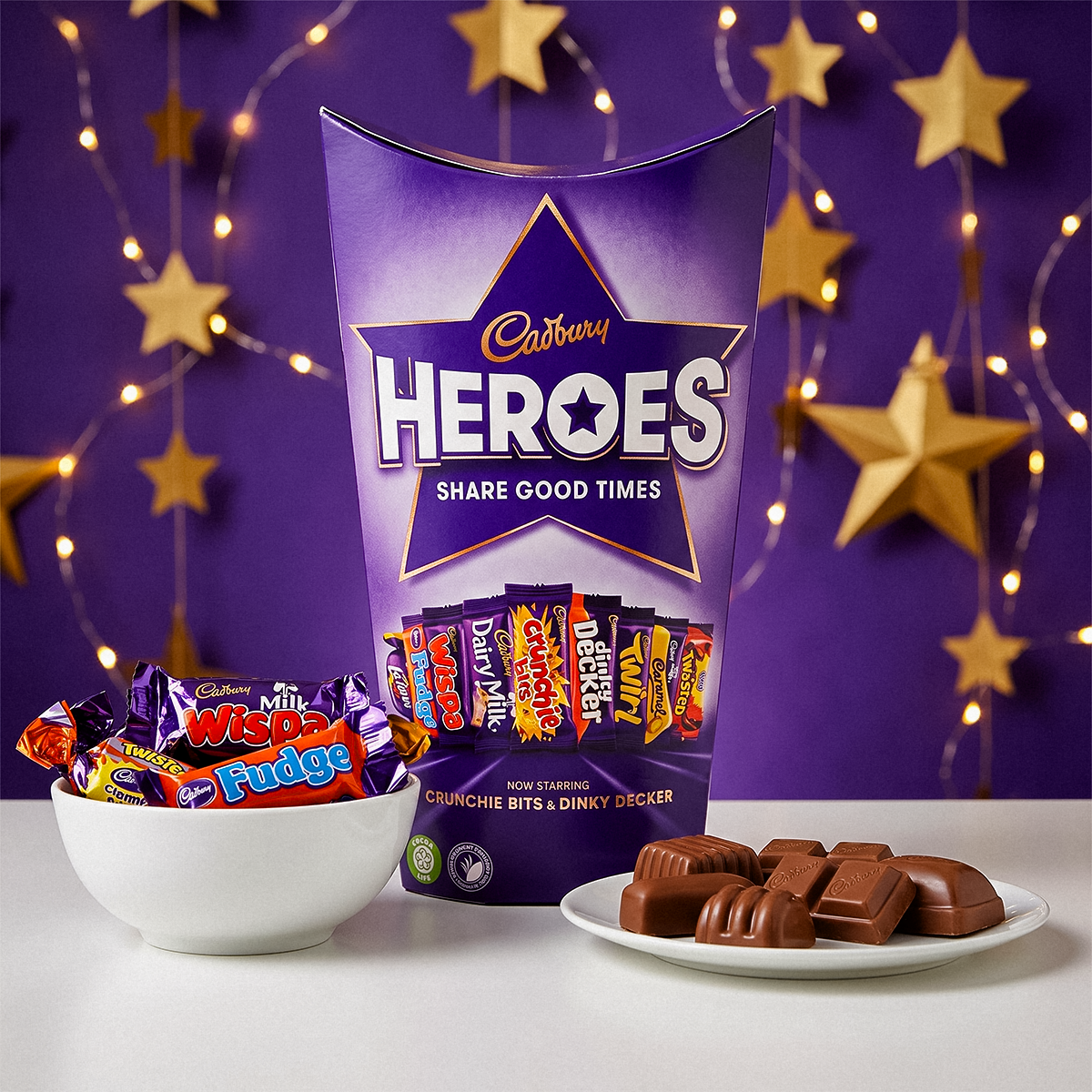 Cadbury Heroes box with a bowl of chocolates, a plate of chocolates, and star garlands on a purple background