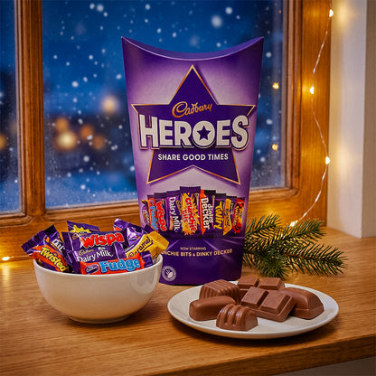 Cadbury Heroes box with a bowl of chocolates, a plate of chocolates, and a snowy window with fairy lights behind