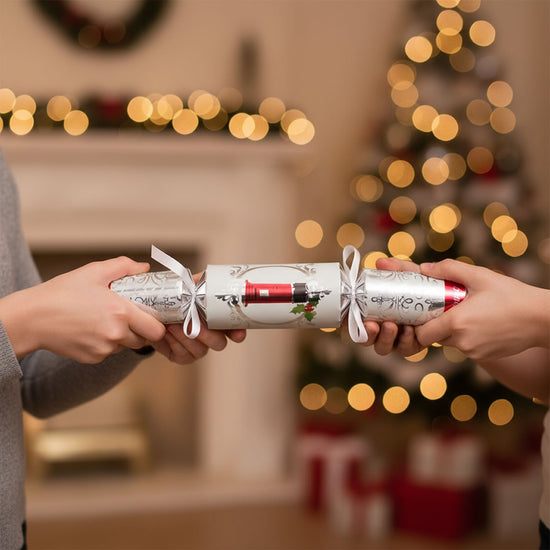 Two hands holding a Christmas cracker in front of a festive background with lights and a fireplace.
