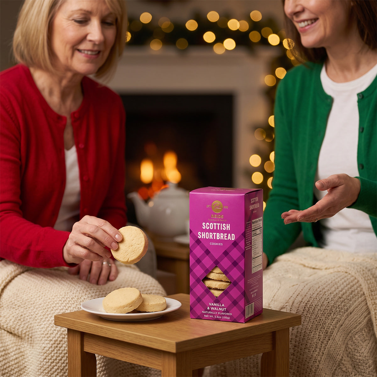 Two older women at a table with a Scottish Shortbread box and a plate of shortbread, fireplace and fairy lights behind