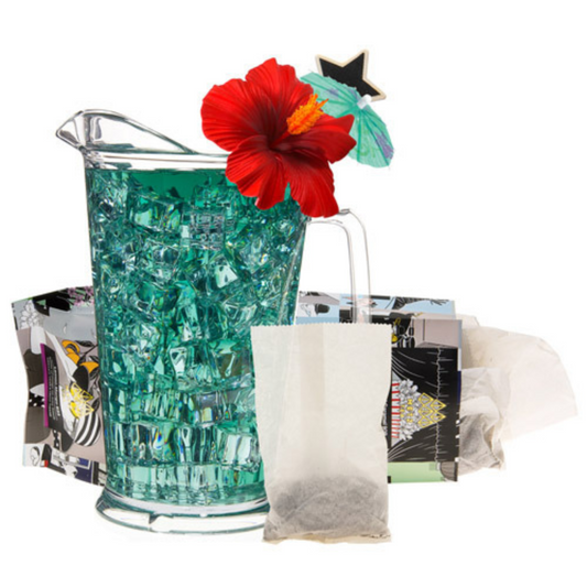 Glass pitcher filled with blue iced tea with ice cubes and a red flower, with tea bags and decorative items in the background.